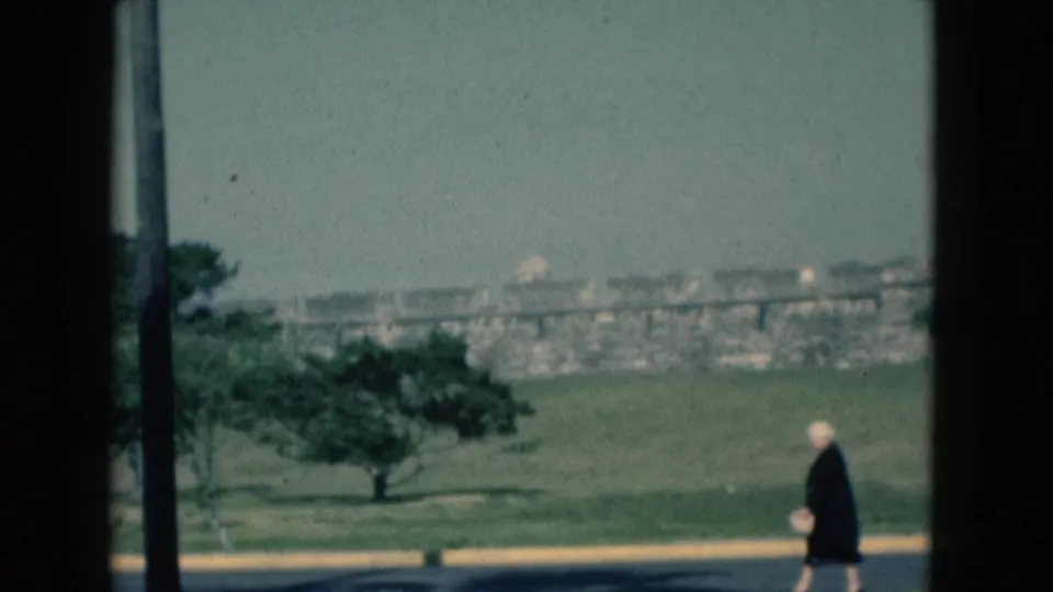 1975 Florida 8mm frame showing an older woman walking beside a road, seen through dark vertical window framing with trees and distant structures.