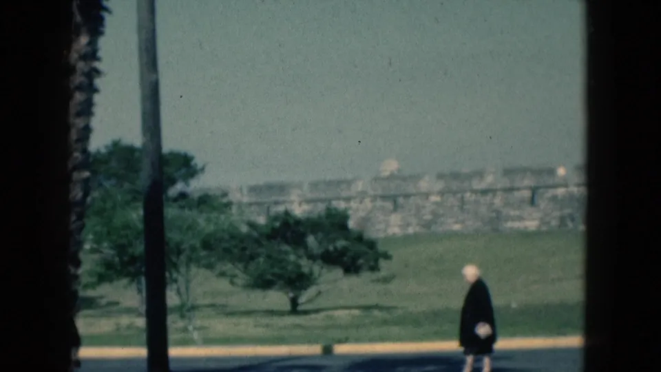 1975 Florida home-movie frame of an older woman walking at the edge of a road, with palm landscaping and distant masonry visible.