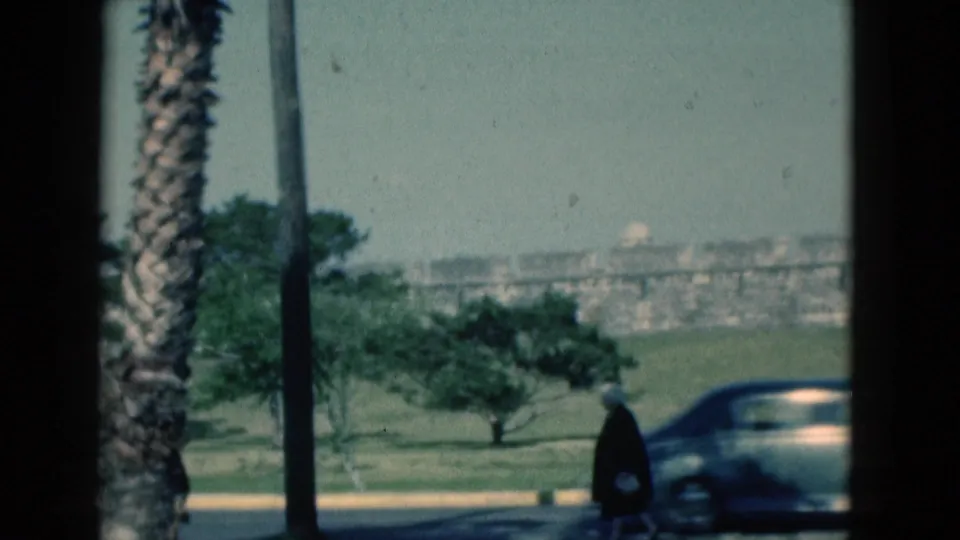 1975 Florida 8mm frame with a classic car passing across the foreground as an older woman walks along the roadside.