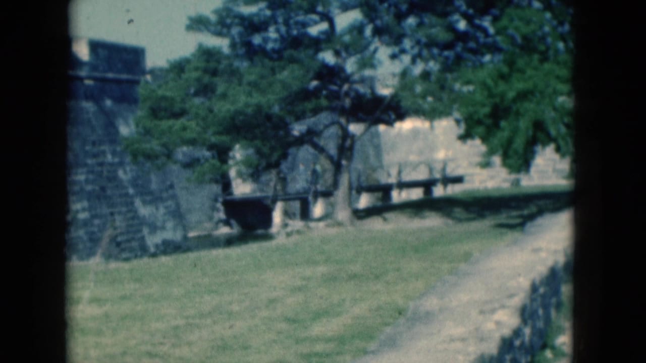 1975 Florida 8mm frame showing a stone fortress wall with grass, trees, benches, and a path in a visitor-shot view.