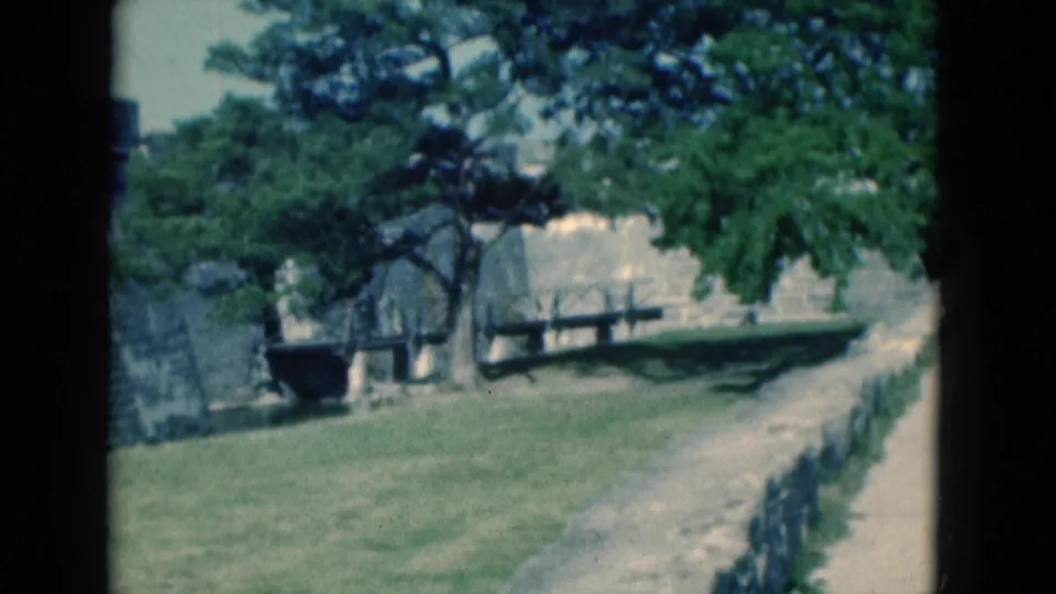 A 1975 Florida home-movie view across tree-shaded fortress grounds with benches, lawn, a path, and stone masonry.