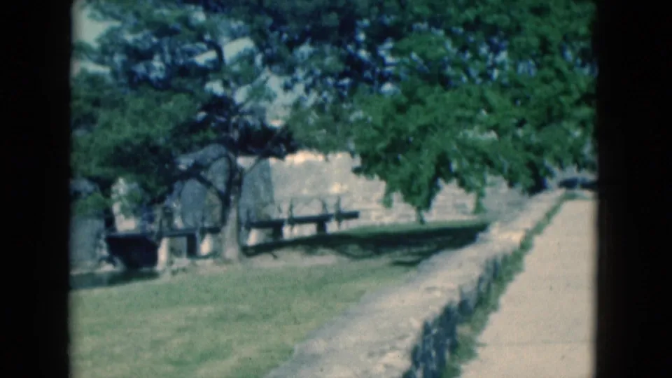 1975 Florida 8mm frame of a walkway running beside lawn, benches, trees, and the edge of a stone fortress wall.