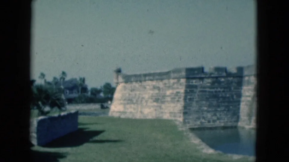 1975 Florida frame showing a stone fortress wall by a moat or water edge, with lawn, trees, and a low wall in front.