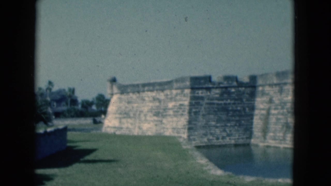 1975 Florida 8mm frame showing a weathered stone fortification, grassy moat edge, still water, and pale sky.