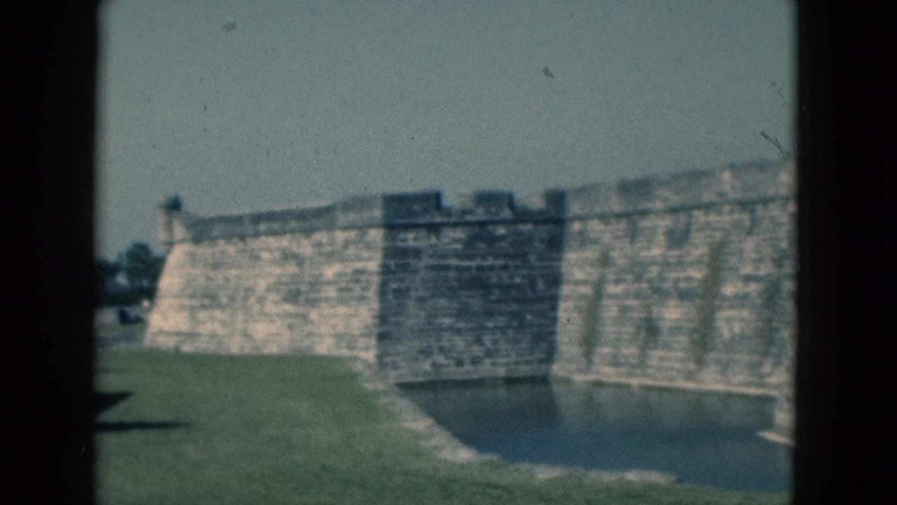 1975 Florida 8mm view of a stone fort wall and surrounding moat, with grass and distant trees under daylight.