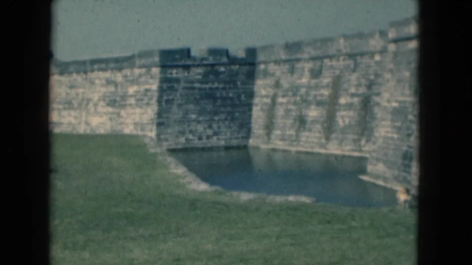 1975 Florida 8mm frame showing the fort wall from across the moat, with grass, water, and soft home-movie color.
