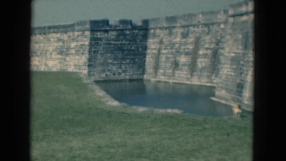 1975 Florida 8mm frame showing a weathered stone fort wall, moat, and grassy foreground from a visitor viewpoint.