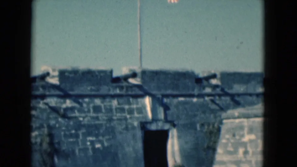 1975 Florida home-movie frame showing cannons on a historic stone fort wall with a flagpole and clear blue sky.