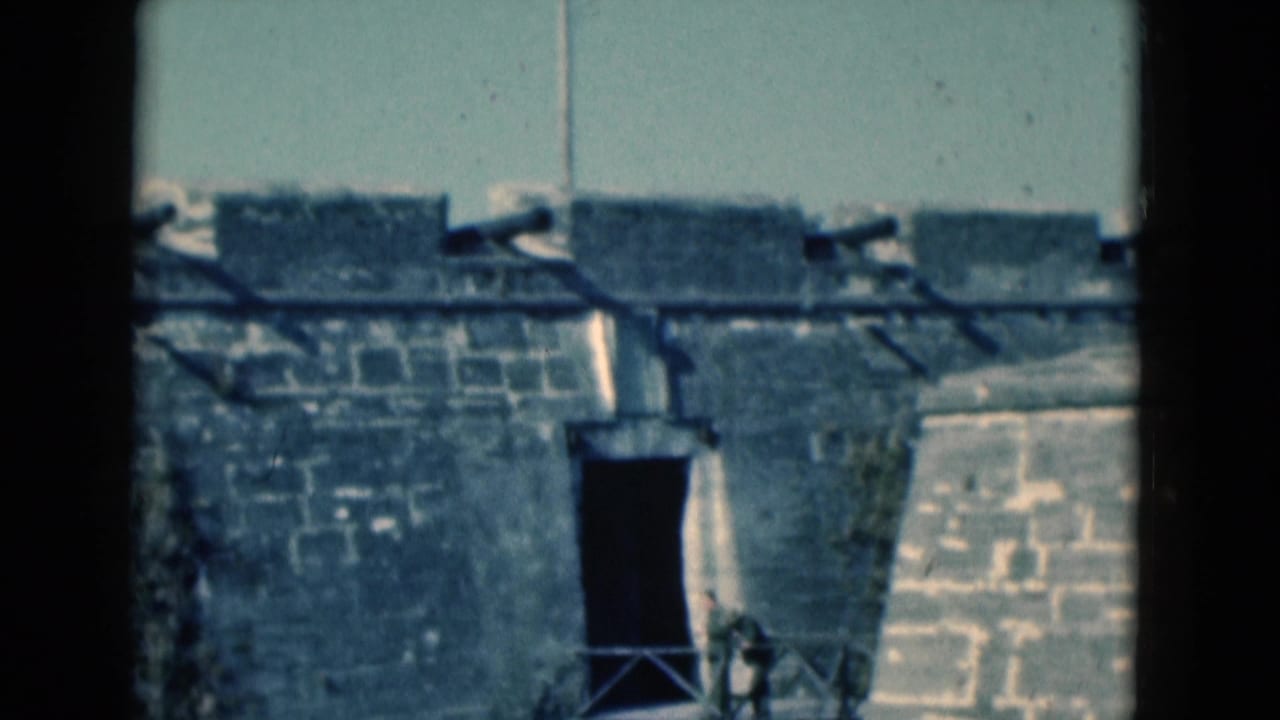 1975 Florida 8mm frame of a stone fort wall with a dark entrance, bridge railing, and water along the base.