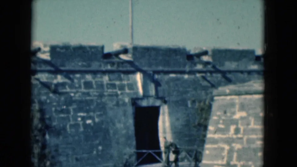 1975 Florida 8mm frame of a stone fort wall with a dark entrance, bridge railing, and water along the base.