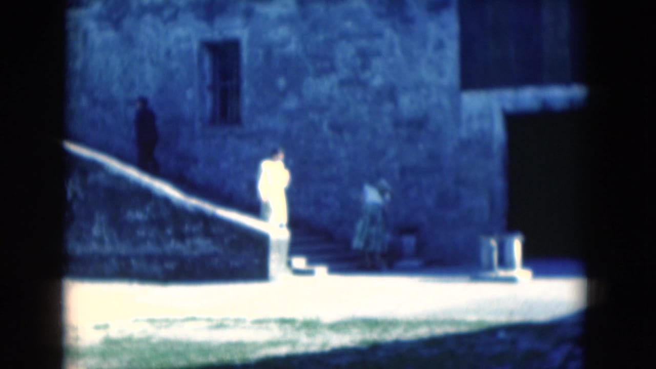 1975 Florida 8mm frame showing a woman in a light dress near stone steps at a weathered stone building, with another figure on the steps.
