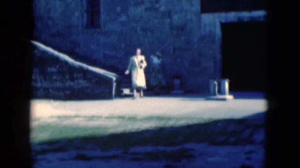 1975 Florida 8mm frame showing a woman at the base of stone steps in a sunlit courtyard beside a weathered stone building.