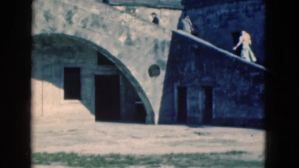 1975 Florida frame showing a stone building with an arched wall, dark doorway, and a woman moving along the upper steps or walkway.