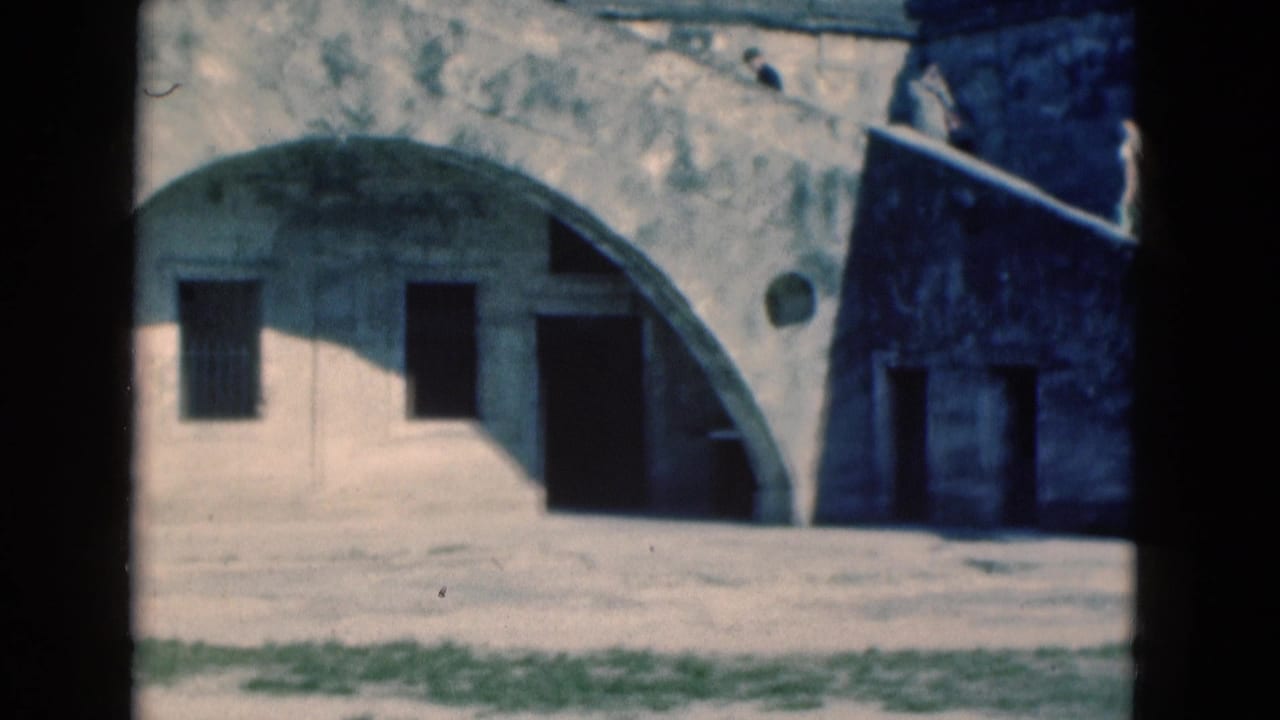 1975 Florida 8mm frame showing a weathered pale building with a large arch, barred windows, and dark door openings.