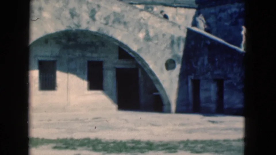 1975 Florida 8mm frame showing a weathered pale building with a large arch, barred windows, and dark door openings.