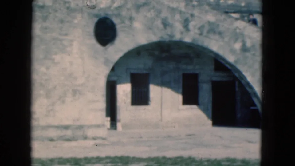 1975 Florida 8mm frame with a closer view of a weathered arched facade, barred window, dark doorway, and open paved foreground.