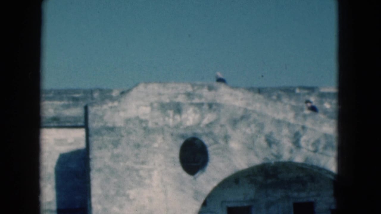 Weathered fort wall with arched openings and small figures along the top under blue sky.