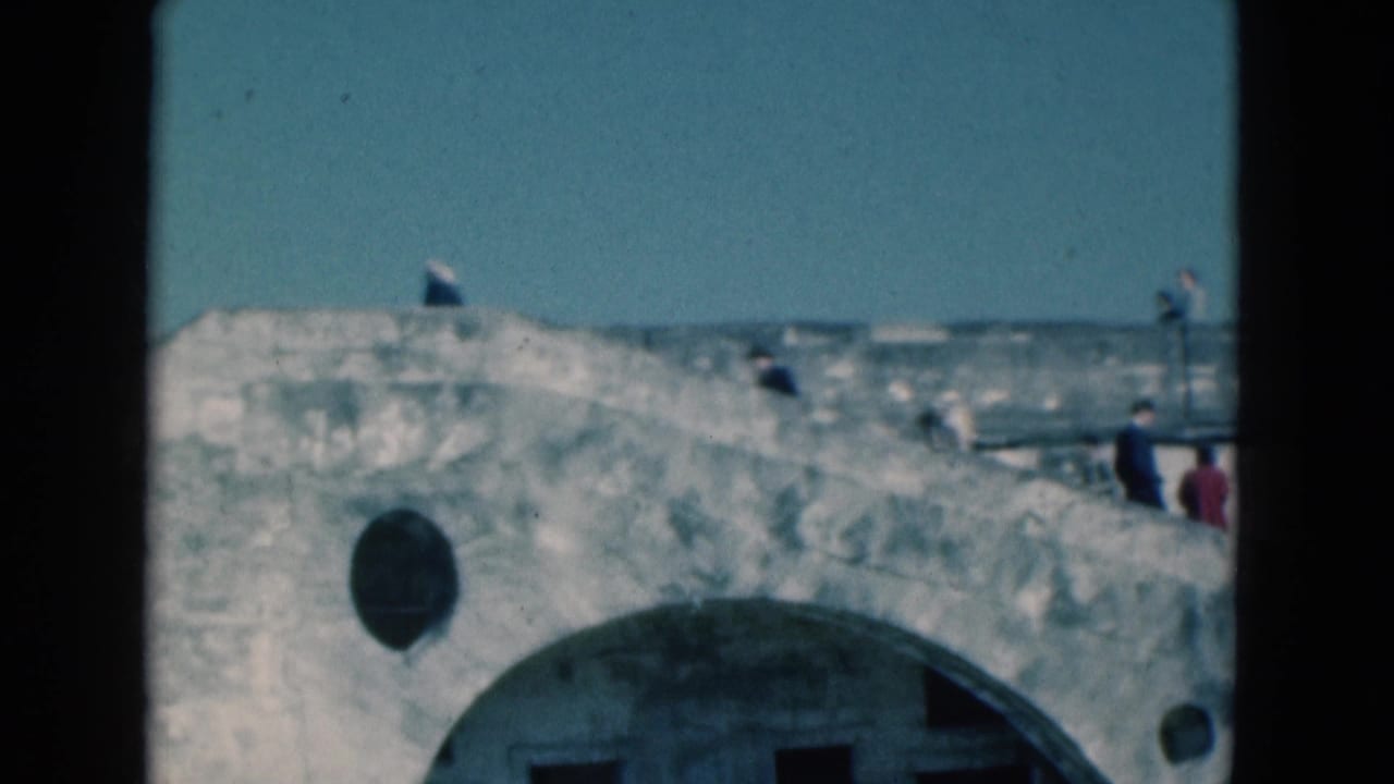 1975 Florida home-movie frame of visitors walking near a weathered fort rampart with an arched opening below.