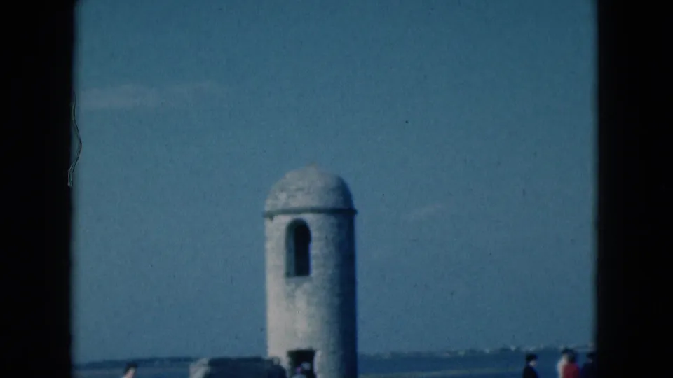 1975 Florida 8mm frame showing a small round fort tower, low wall, distant water, and visitors at the edge of the scene.