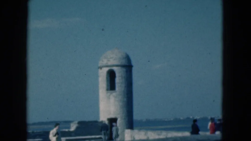 1975 Florida home-movie frame of a round fort tower beside low ramparts, with visitors walking under a clear blue sky.