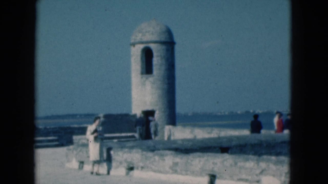 1975 Florida home-movie frame of a woman in a light dress walking past a stone walkway with the ocean beyond.