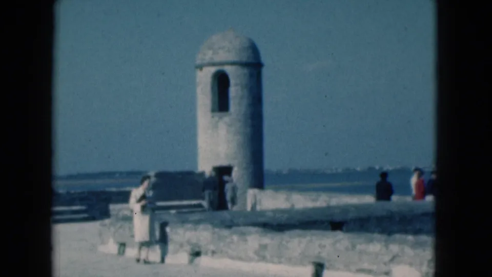 1975 Florida home-movie frame of a woman in a light dress walking past a stone walkway with the ocean beyond.
