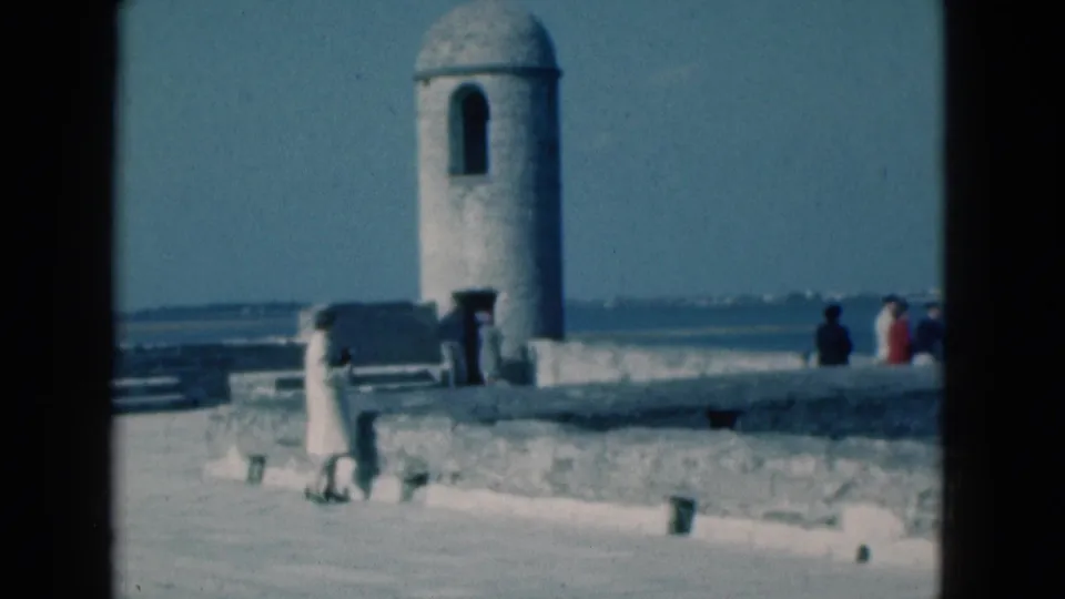 1975 Florida 8mm frame with a woman walking in front of a stone parapet while visitors stand near the sentry tower.