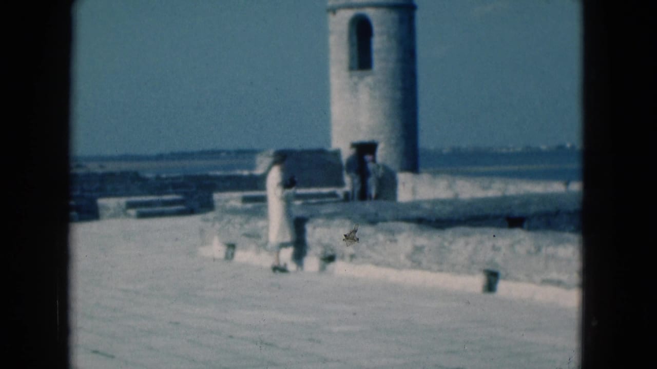 Person in a light dress stands on a stone waterfront near a cylindrical tower and blue water.