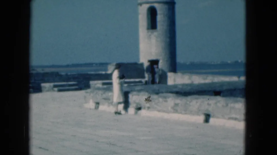 1975 Florida 8mm frame showing a person in a light dress near a stone pier, with a cylindrical tower and calm water behind them.