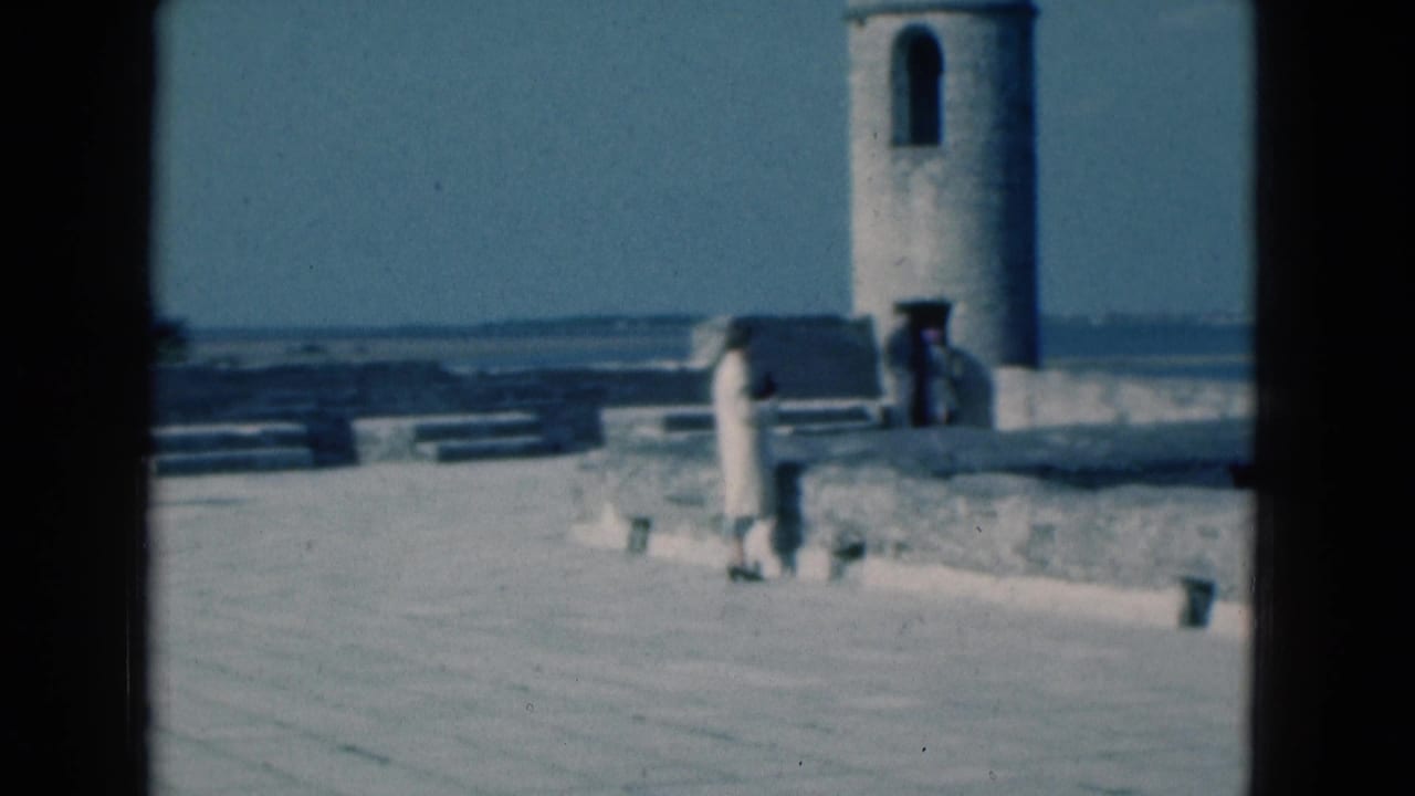 1975 Florida coastal view with a distant person on a stone pier, benches or steps nearby, and a round tower beside calm water.