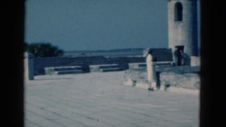 Soft 8mm view of a Florida waterfront terrace in 1975, with a person near the stone wall and a tower rising at the right edge.