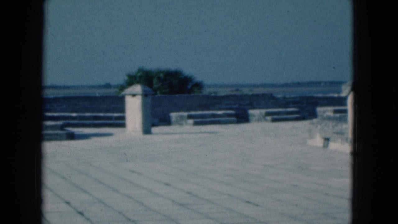 1975 Florida home-movie view of a sunlit stone patio, low walls, distant water, and a small white tower framed by dark mullions.