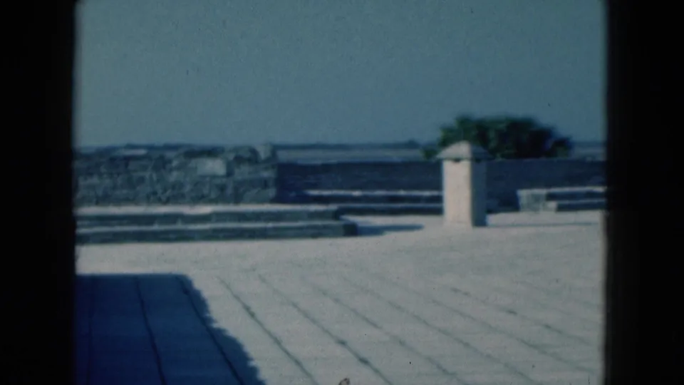 1975 Florida terrace scene with stone paving, a small tower, waterfront wall, and pronounced window-frame borders from the camera position.