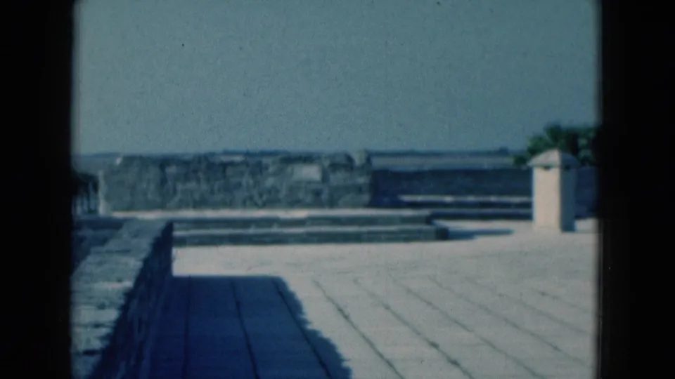 Final 1975 Florida home-movie frame showing the stone patio, low seawall, distant water, and small white tower through a window.
