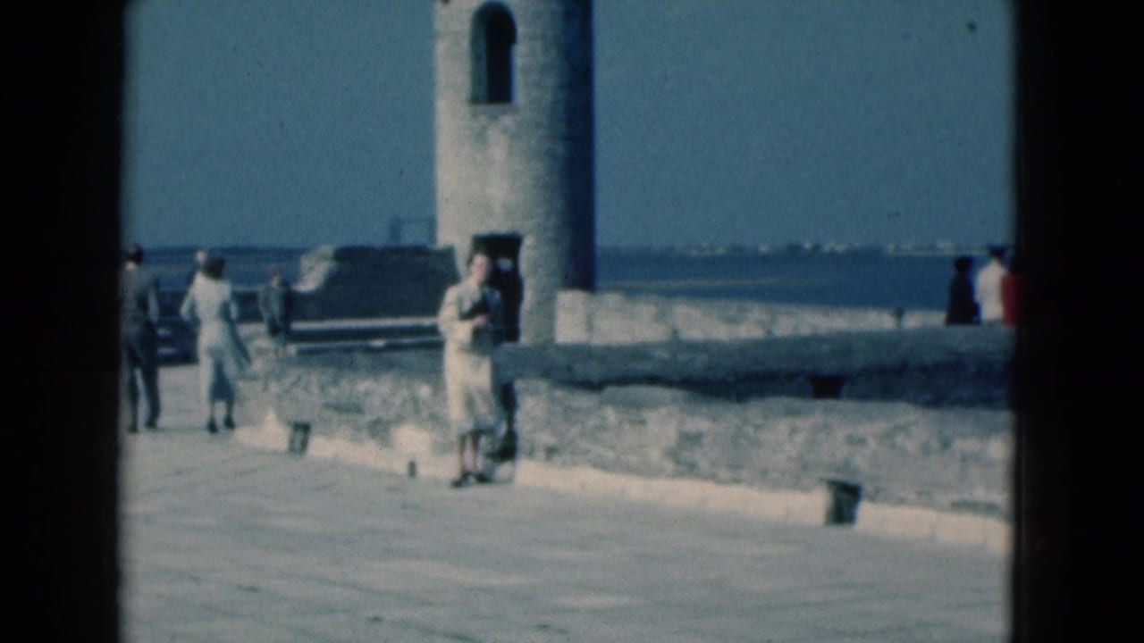 1975 Florida 8mm frame showing a woman on a waterfront promenade near a stone tower, with visitors and water behind her.