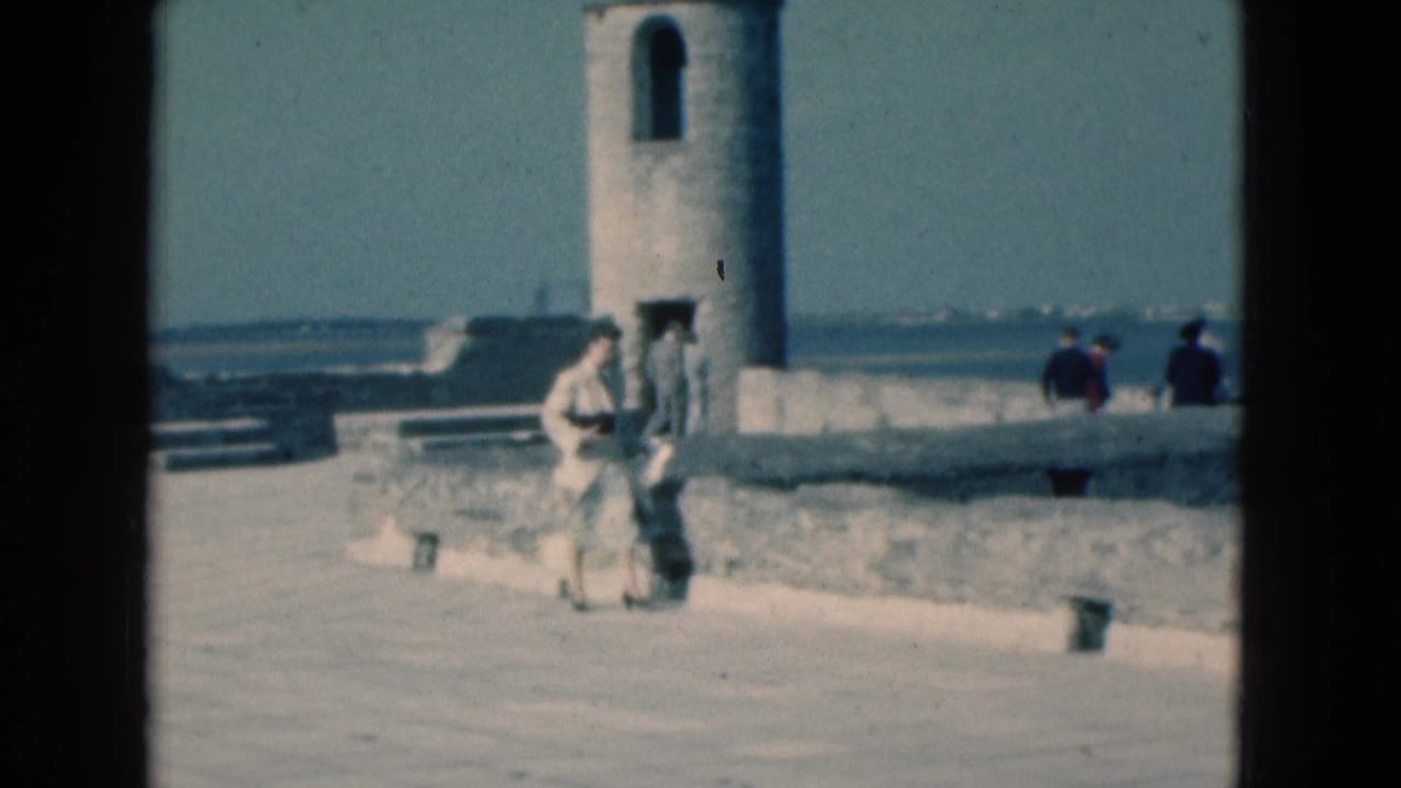 Woman standing near the promenade wall with a child-sized figure beside her and a stone tower behind.