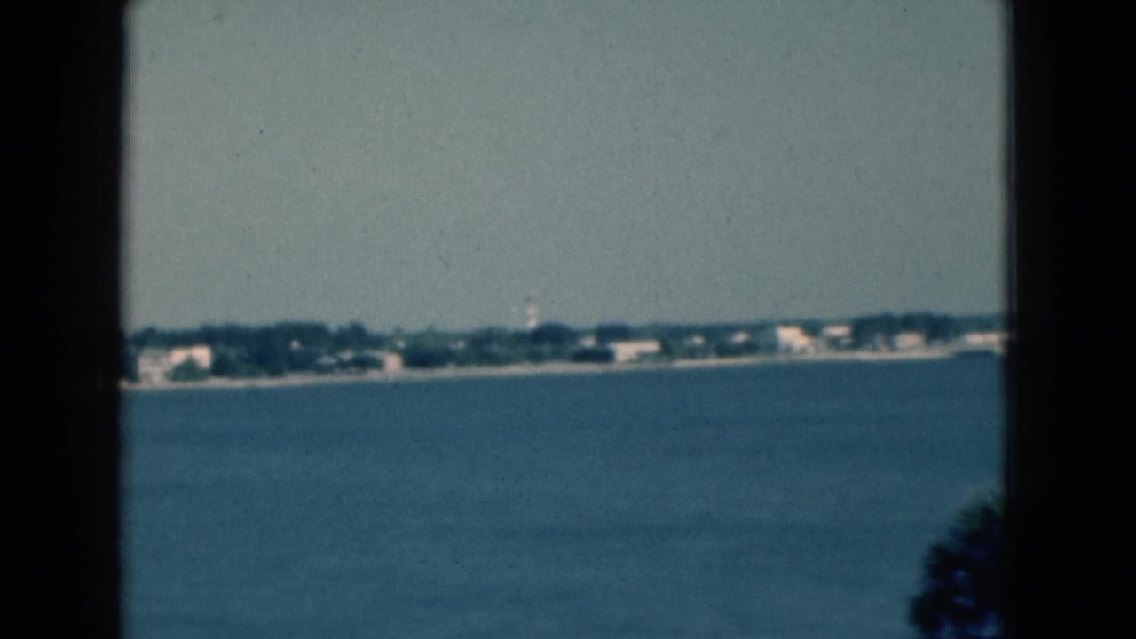 1975 Florida 8mm frame showing blue water, a distant shoreline, low buildings, trees, and dark window framing in the foreground.