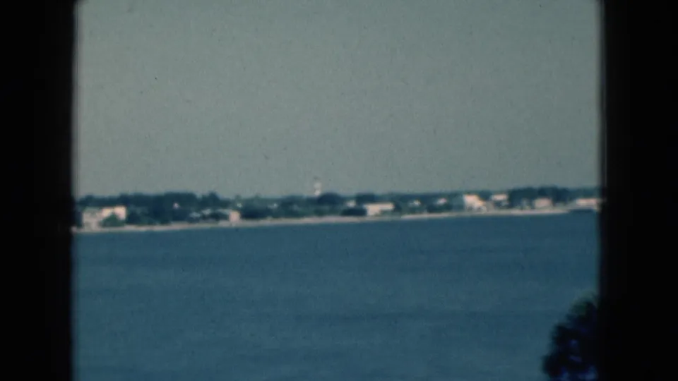 1975 Florida 8mm frame showing blue water, a distant shoreline, low buildings, trees, and dark window framing in the foreground.