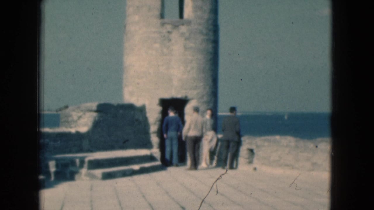 1975 Florida 8mm frame showing adult tourists gathered by a stone tower doorway on a seaside ruin platform.