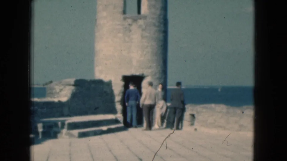 1975 Florida 8mm frame showing adult tourists gathered by a stone tower doorway on a seaside ruin platform.