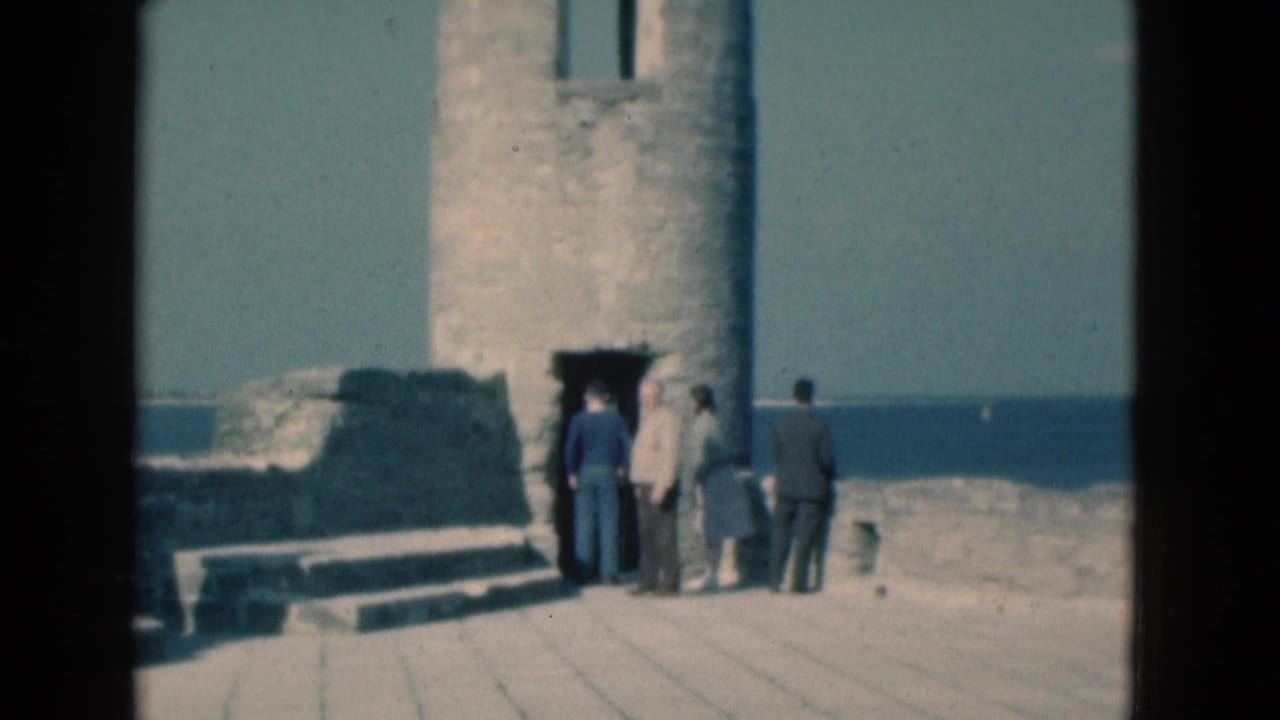 A small group of adults stands beside a stone tower doorway on a sunlit coastal platform.