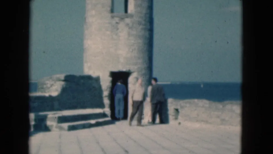 1975 Florida home-movie frame of adult tourists at a weathered seaside tower ruin, seen from a handheld 8mm perspective.