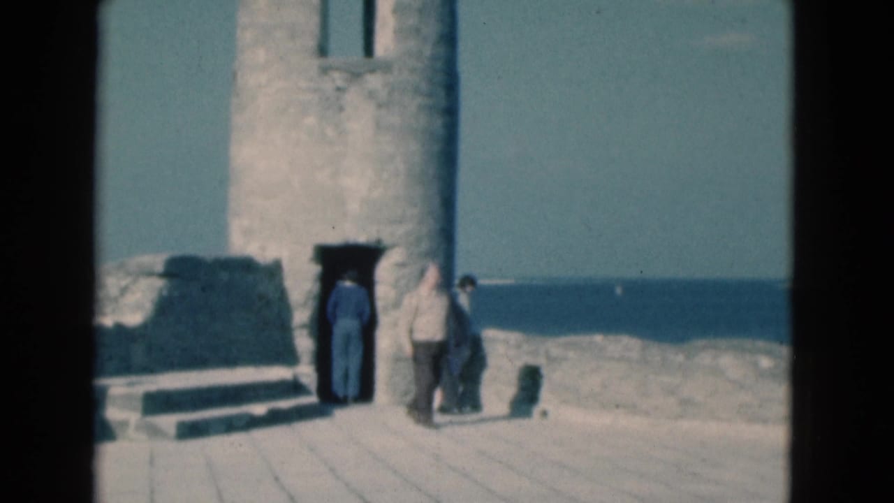 1975 Florida home-movie frame showing adults near a weathered stone lighthouse doorway with ocean beyond.