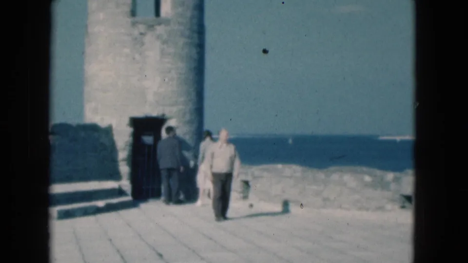 1975 Florida home-movie still of adults walking past a lighthouse doorway on a sunlit coastal platform.