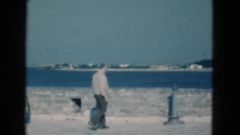 1975 Florida coastal frame showing an adult man walking past a seawall with ocean and distant shoreline beyond.