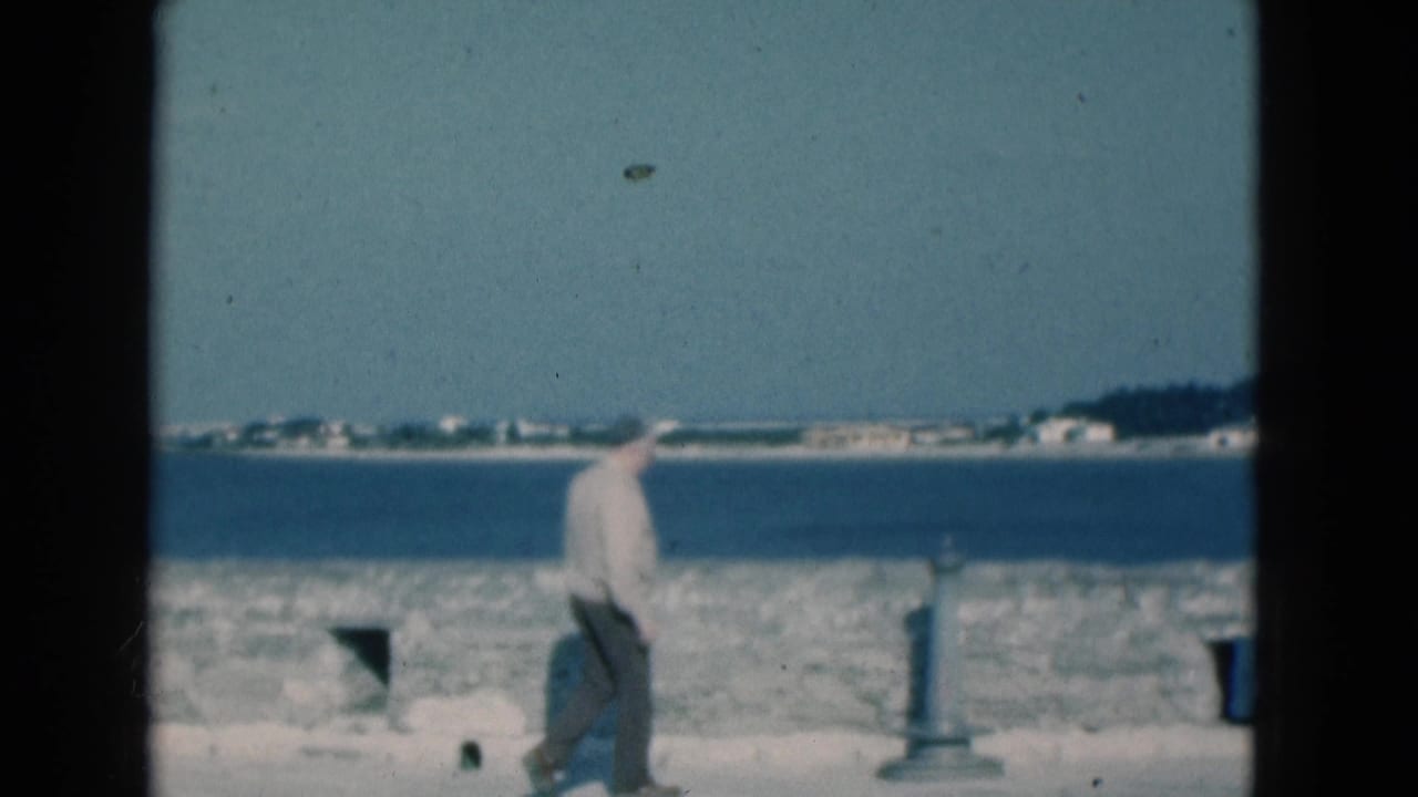 Man walking beside a stone seawall with blue water and distant shoreline behind him.
