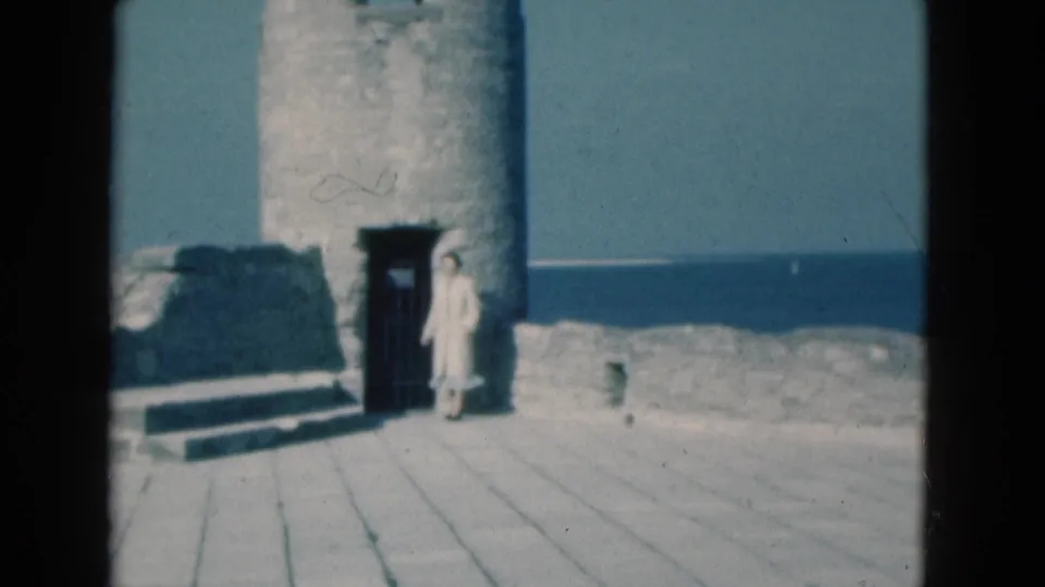 1975 Florida frame showing a person near a round stone tower, doorway, paved walk, and water beyond.
