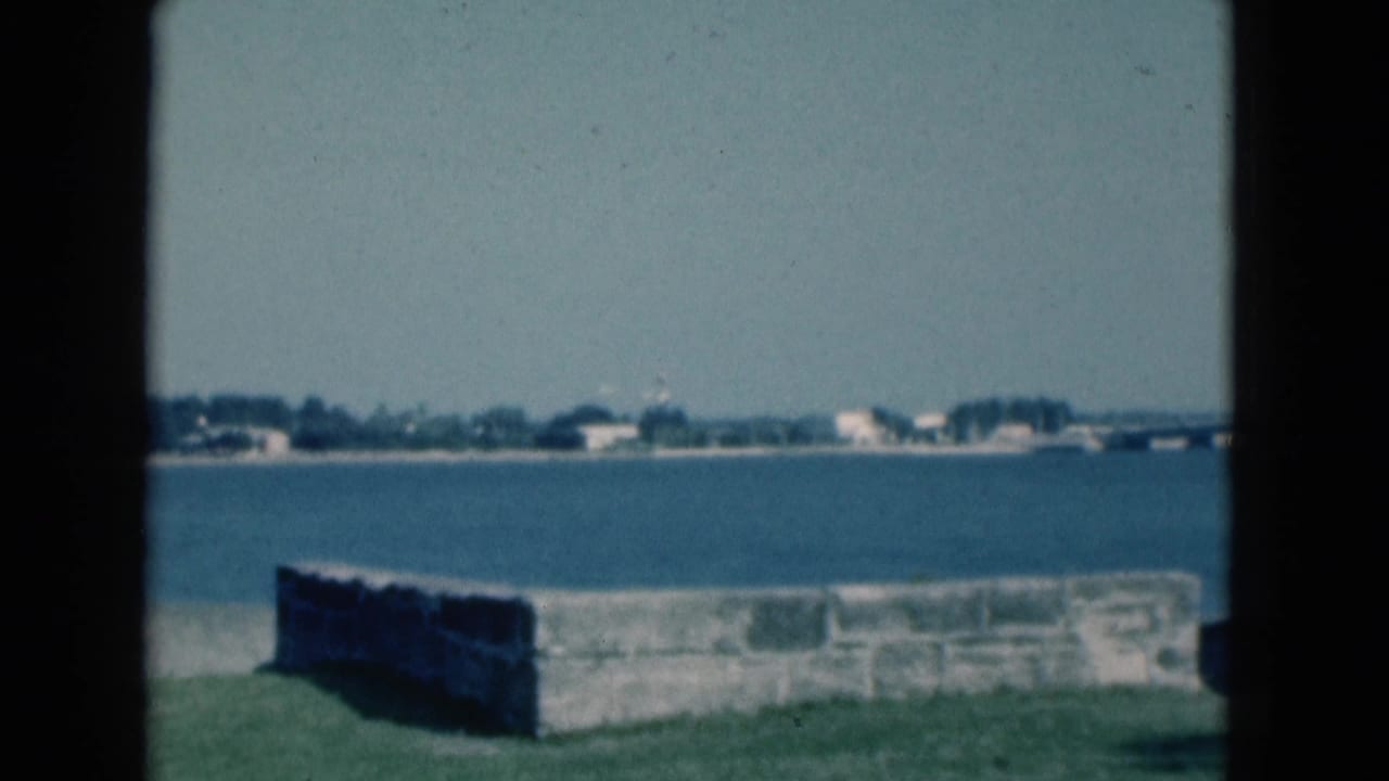 Grassy park edge with a low stone wall beside blue water and a distant shoreline.