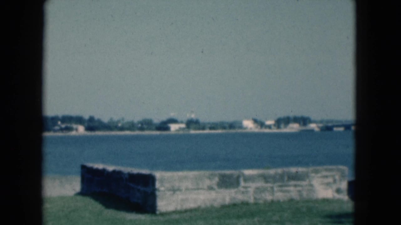 Static 1975 Florida coastal view from a park, with a masonry wall, calm blue water, and pale shoreline buildings in the distance.
