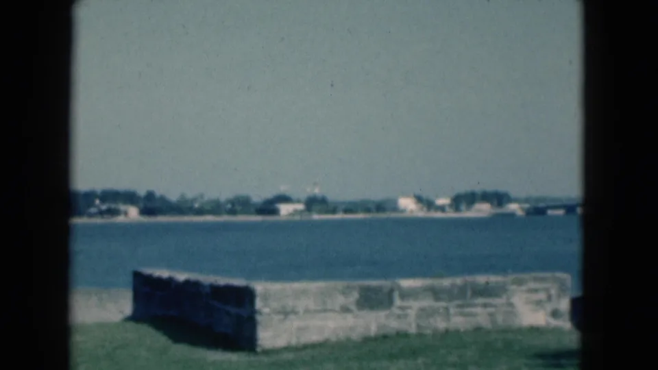 Static 1975 Florida coastal view from a park, with a masonry wall, calm blue water, and pale shoreline buildings in the distance.